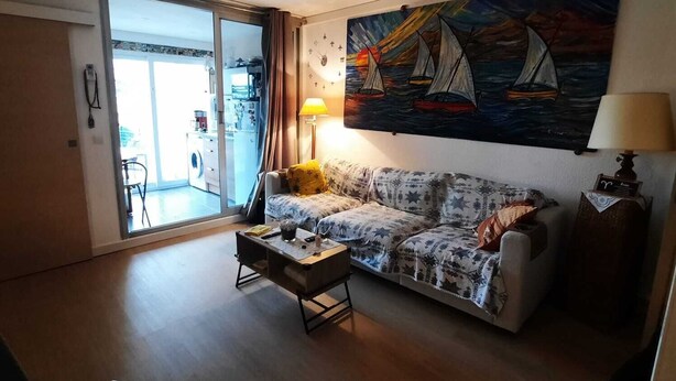 Ein Wohnzimmer mit einer Couch, einem Couchtisch, einem Gemälde mit Segelbooten, einer Lampe und einer Glastür zum Balkon.