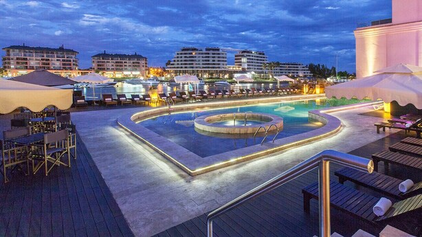 A rooftop pool area with lounge chairs, a hot tub, and a view of surrounding buildings at dusk.