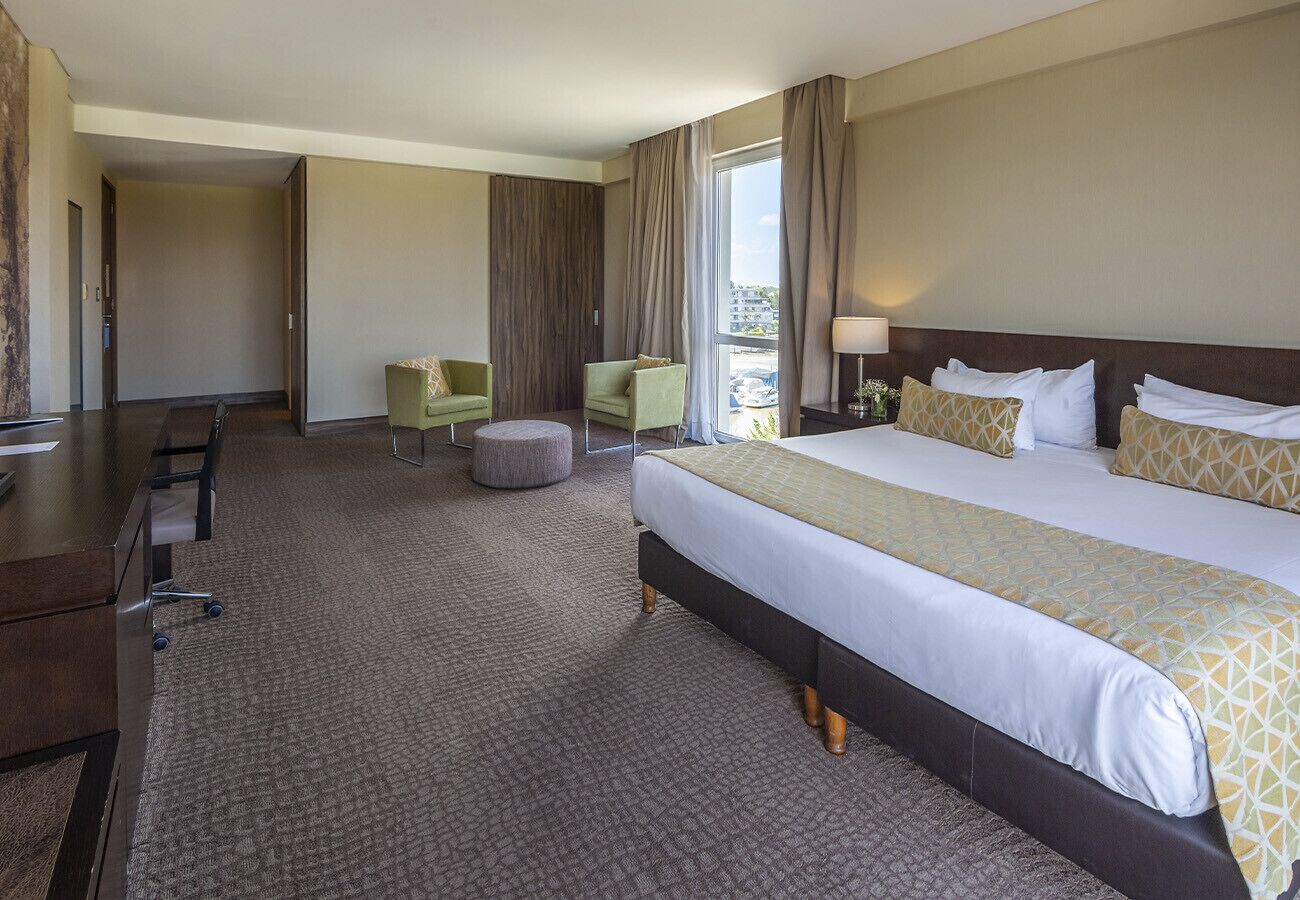 A hotel room with a large bed, a desk, two chairs, a small ottoman, and a view of the city.