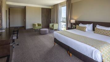 A hotel room with a large bed, a desk, two chairs, a small ottoman, and a view of the city.