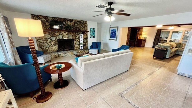A living room with a stone fireplace, a sofa, a blue armchair, a wooden floor lamp, and a ceiling fan.