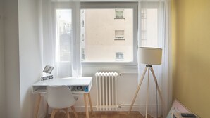 Et værelse med et hvidt skrivebord, en stol, en radiator, en gulvlampe og et vindue med udsigt til bygninger.
