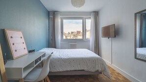 Et soveværelse med seng, skrivebord, stol, et vindue med gardiner, en radiator og et tv ophængt på væggen.