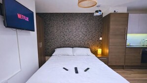 Een slaapkamer met een bed, een televisie aan de muur met 'NETFLIX', een klein nachtkastje met een lamp en een kitchenette met kastjes.