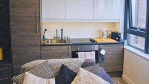 Een kleine keuken met witte kastjes, een zwart aanrecht, een raam en een bed met kussens met een patroon.