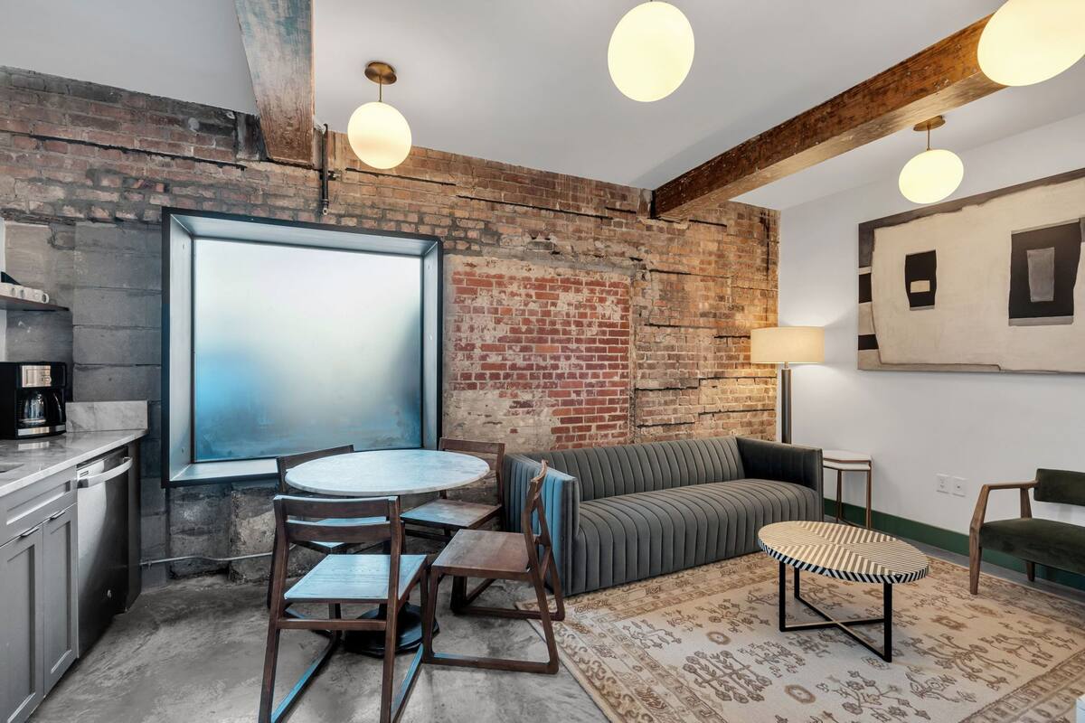 A room with a brick wall, a round table with chairs, a sofa, a patterned rug, and pendant lighting.