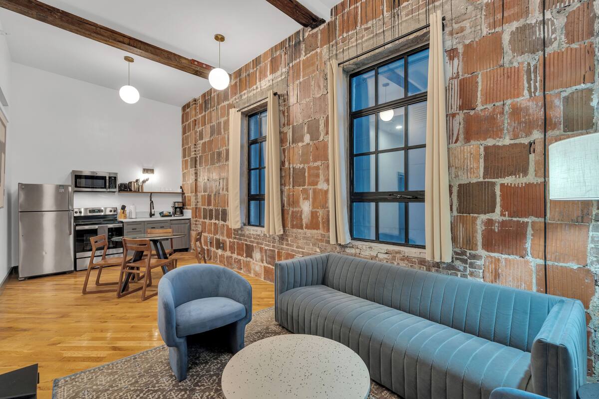 A modern kitchen with stainless steel appliances, a dining area with wooden chairs, and a large brick wall with windows.