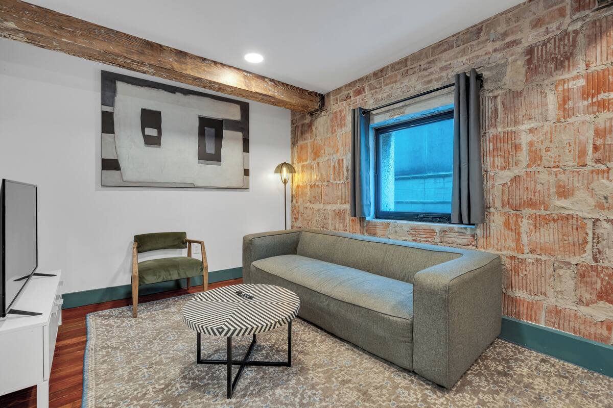 A room with a brick wall, a window, a sofa, a chair, a small table, and a lamp.