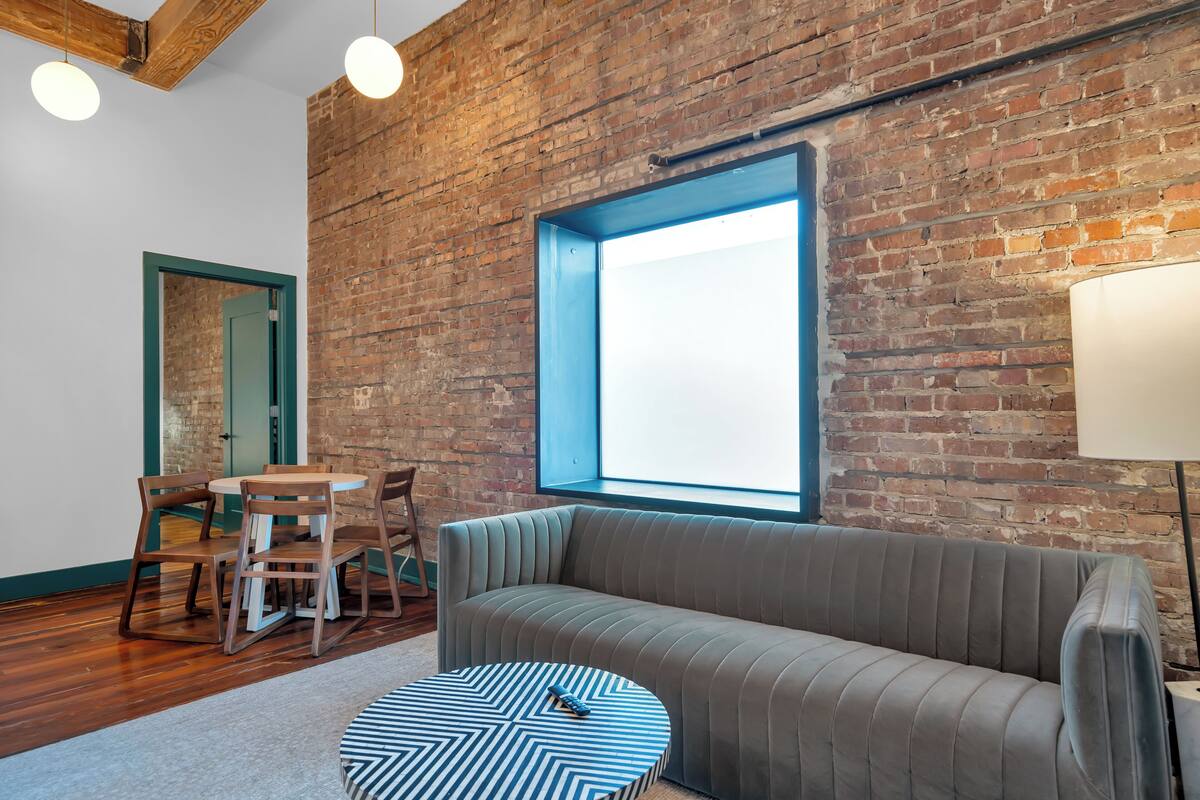 A room with a brick wall, a large window, a sofa, a round table, and a dining area with chairs.