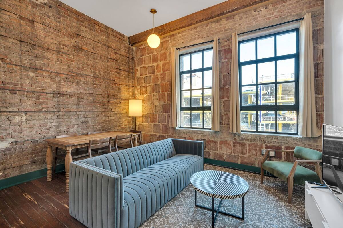 A room with a brick wall, a sofa, a dining table, a chair, a lamp, and a TV.