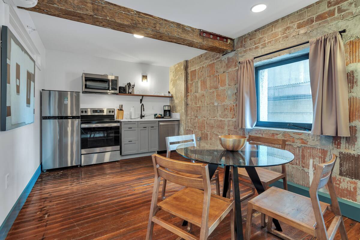 A kitchen with stainless steel appliances, a brick wall, and a dining area with a glass table and wooden chairs.