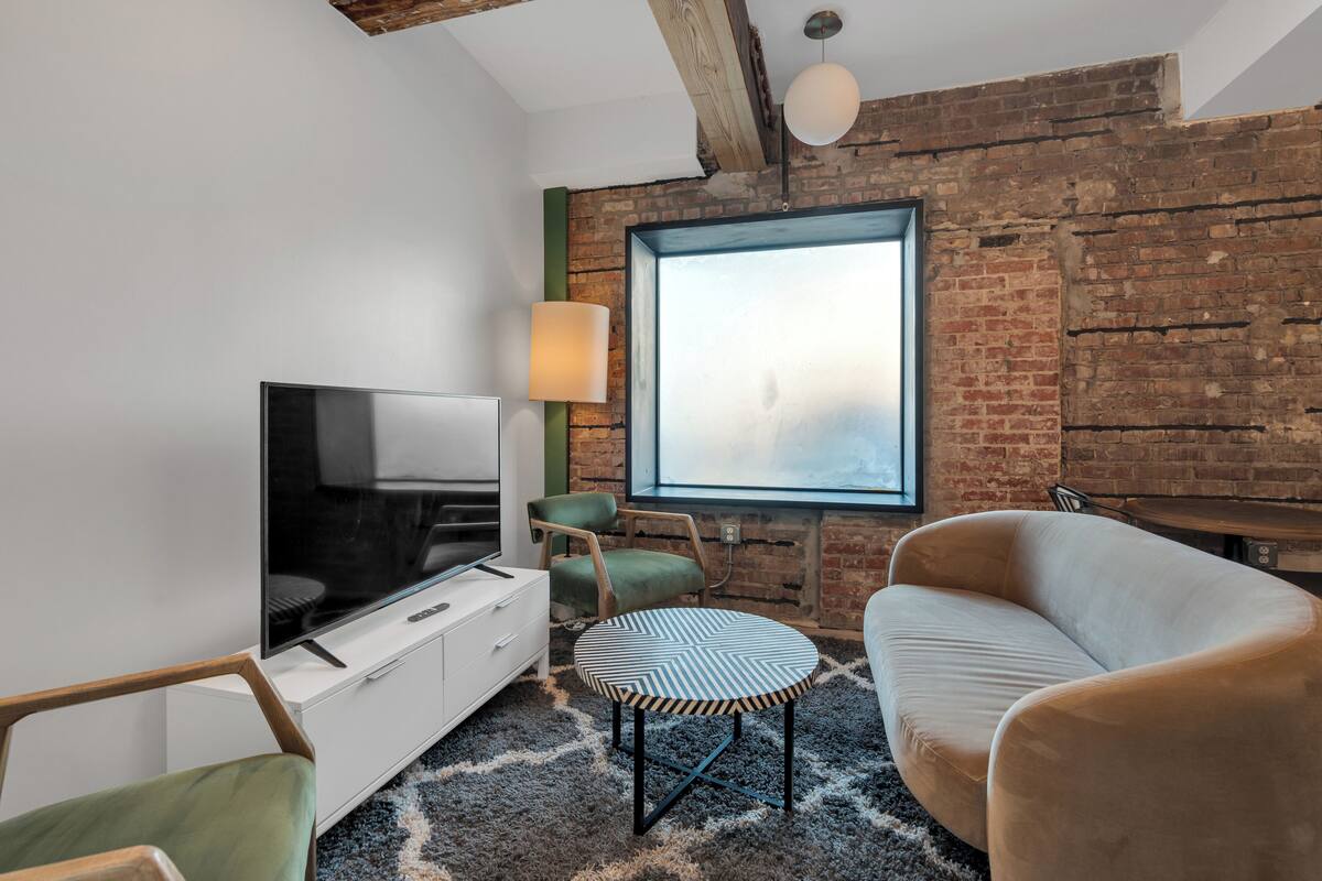 A modern living room with a television, seating, a large window, and exposed brick walls.