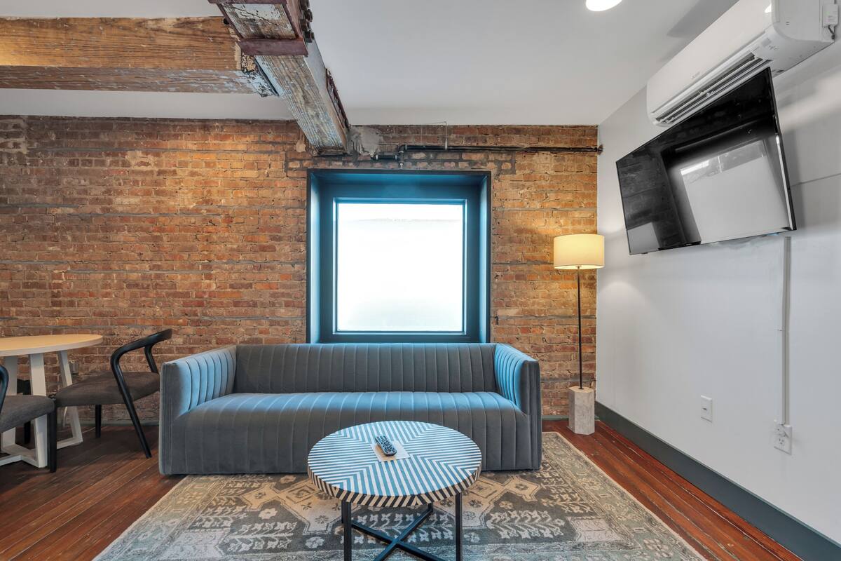 A room with a brick wall, a blue-framed window, a gray sofa, a round table, and a mounted TV.
