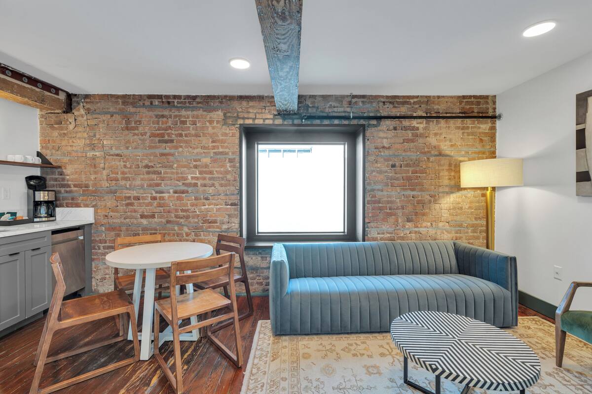 A room with a brick wall, a window, a sofa, a table with chairs, and a lamp.