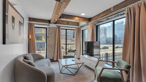 A modern living room with a sofa, a coffee table, and a TV. There are large windows with curtains, a view of the city, and a balcony. - Schaeffer Hotel (New Orleans)