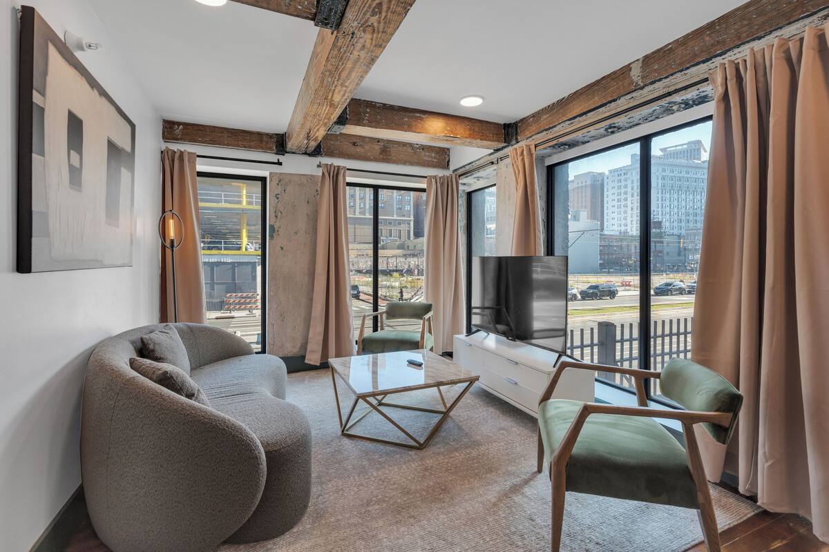 A modern living room with a sofa, a coffee table, and a TV. There are large windows with curtains, a view of the city, and a balcony.