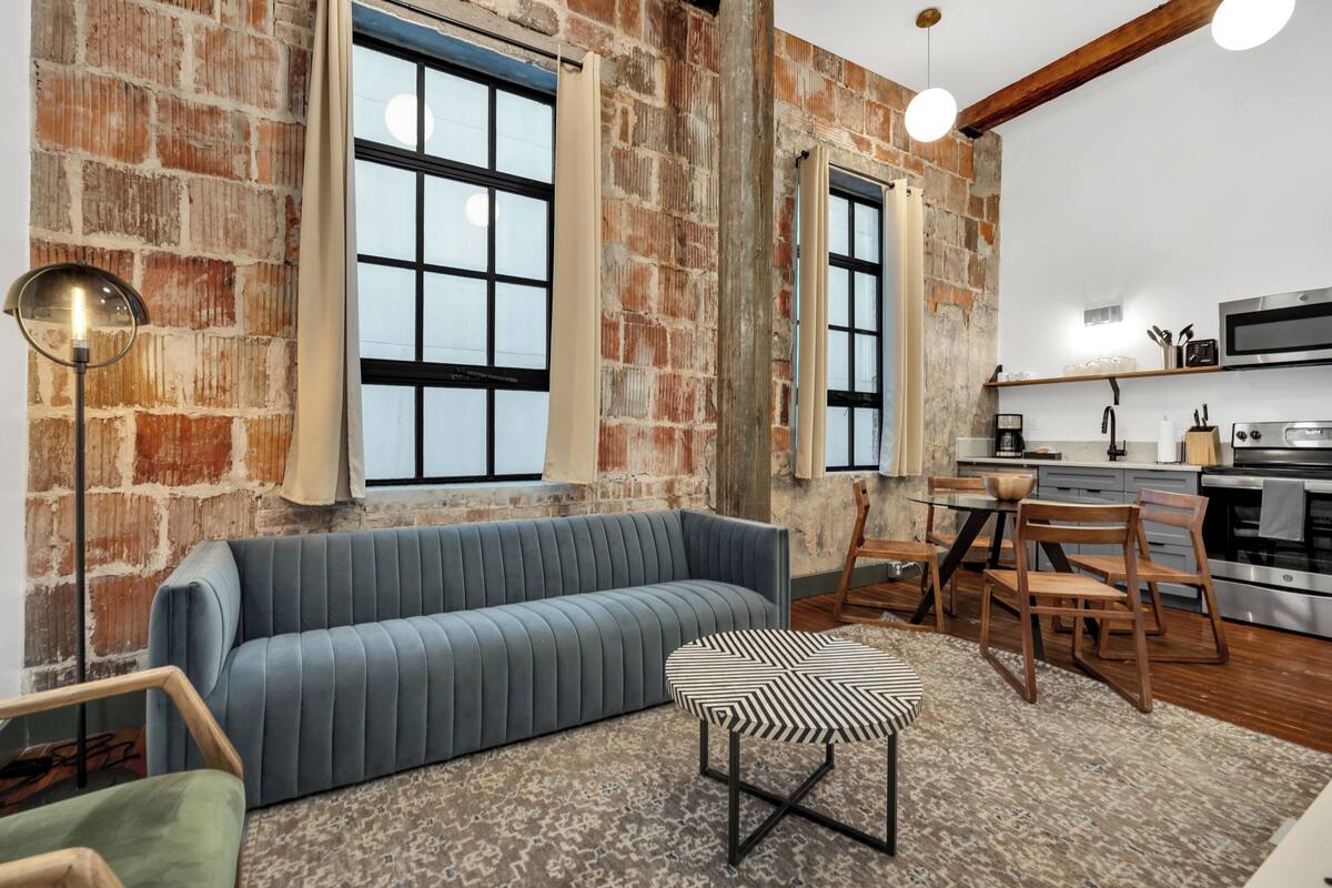 A modern kitchen with a built-in oven, a dining area with a table and chairs, and a blue sofa in a room with exposed brick walls and large windows.