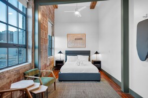 Een moderne slaapkamer met een groot bed, twee nachtkastjes, een plafondventilator, uitzicht op de stad en een bakstenen muur.
