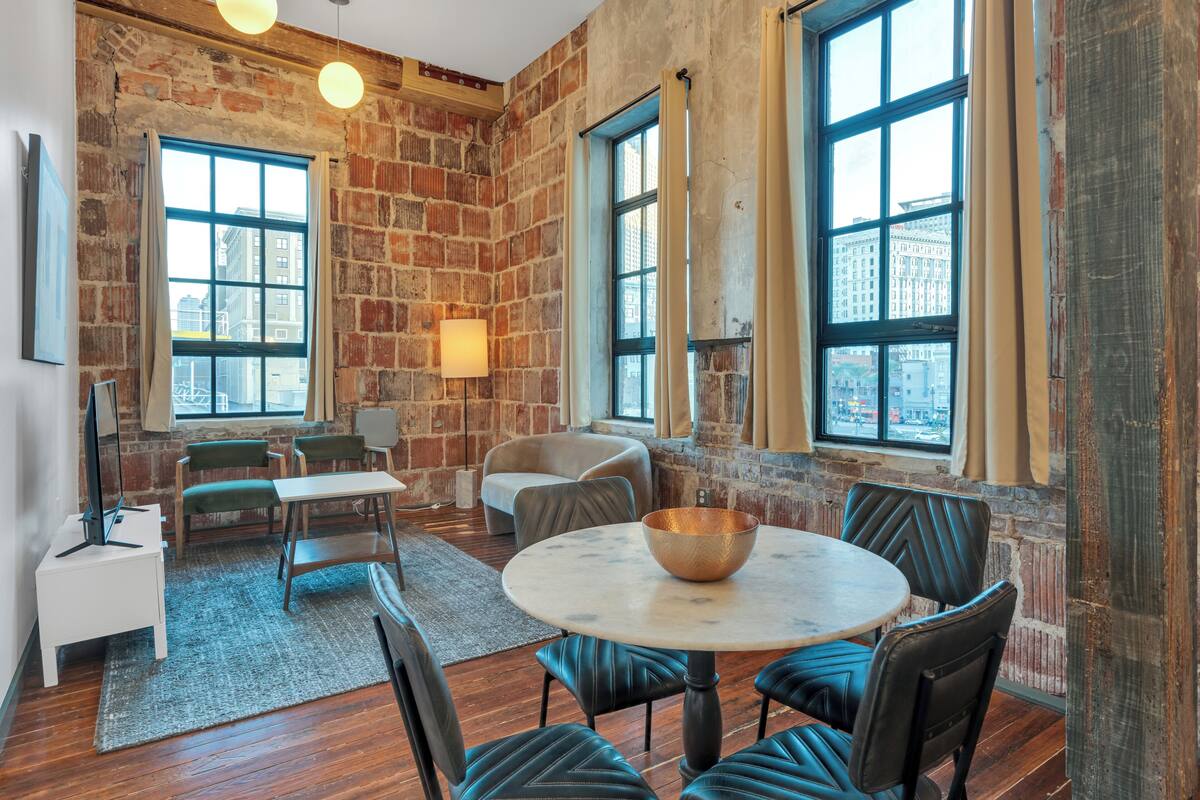A room with a brick wall, large windows, a round table with a bowl, and leather chairs.