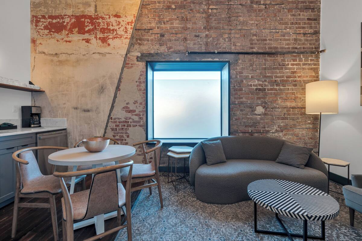 A loft-style room with a brick wall, a large window, a sofa, a round dining table with chairs, and a small side table.