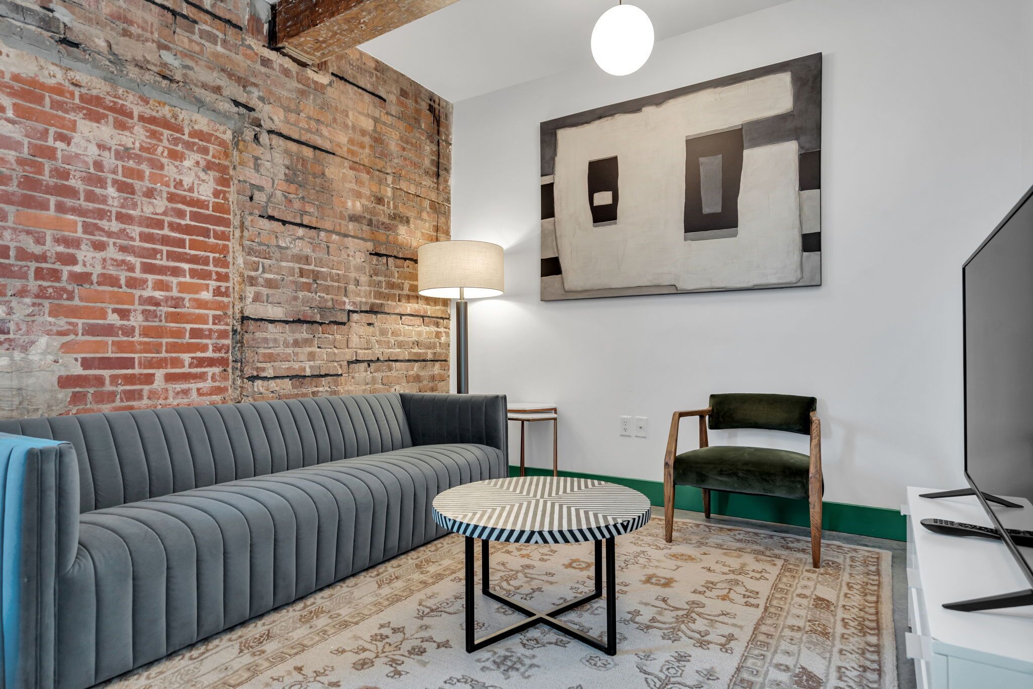 A room with a brick wall, a grey sofa, a round table, a lamp, a chair, and a TV.
