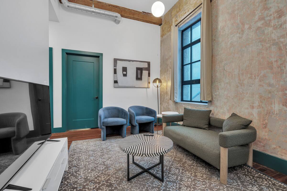 A modern living room with a teal door, two blue armchairs, a gray sofa, a patterned coffee table, and a large window.