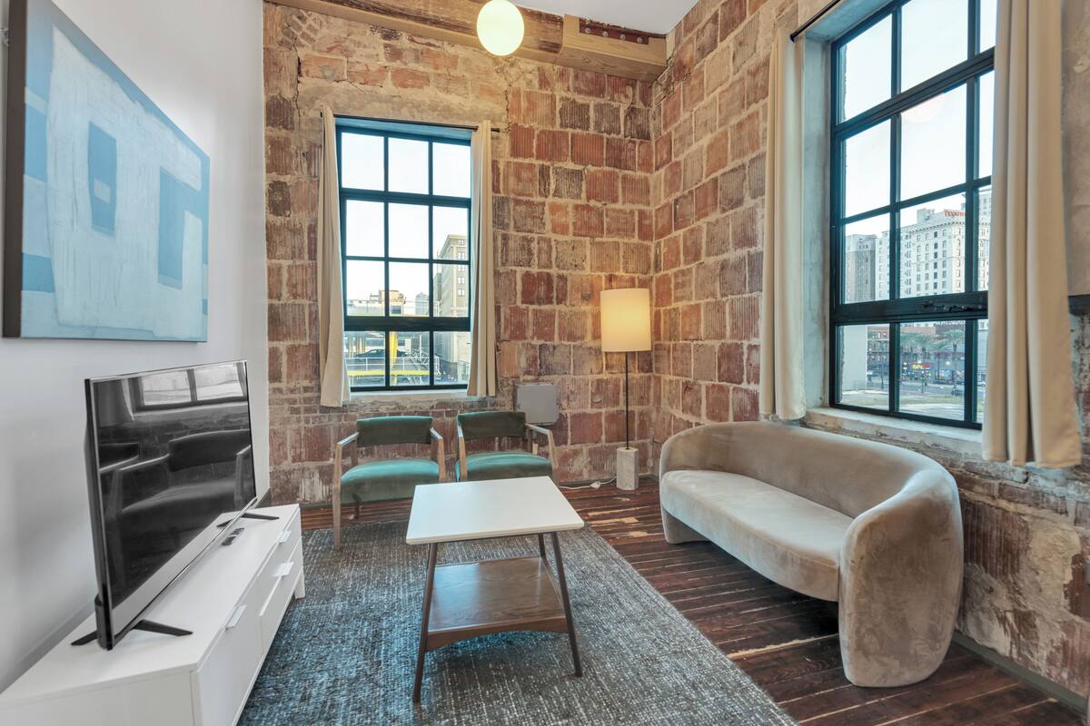 A room with a brick wall, a large window, a sofa, a TV, and a table.