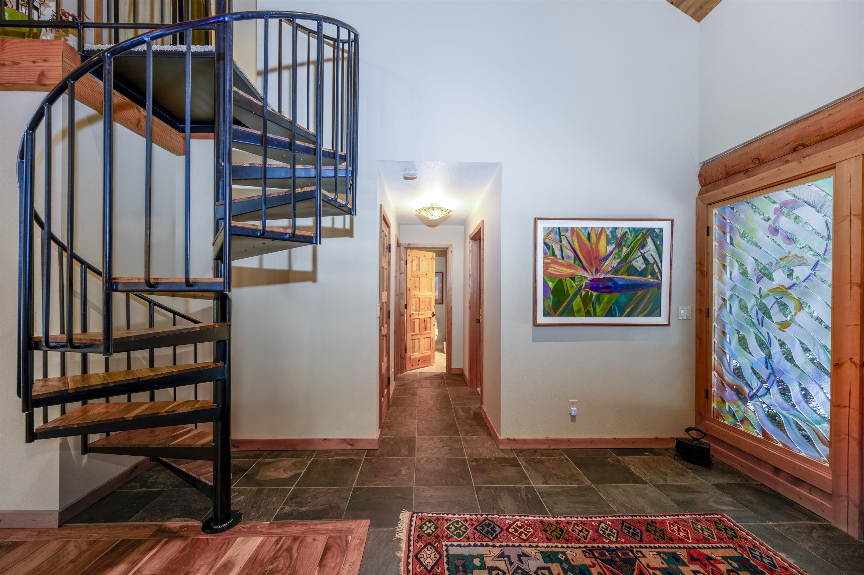 Un escalier en colimaçon avec des marches en bois et des rampes en métal, menant à un couloir orné d’un tapis à motifs et d’une peinture abstraite aux couleurs vives accrochée au mur.