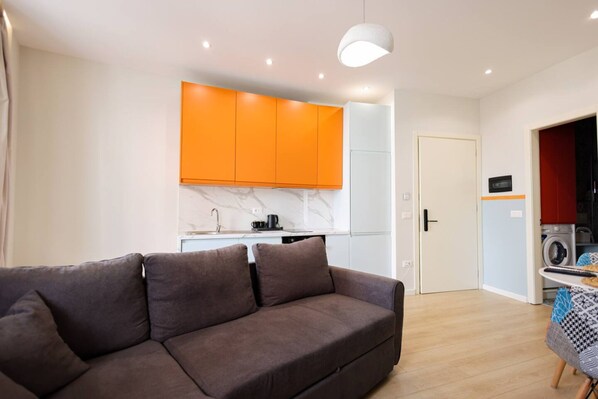 A modern living room with a sofa, a kitchen with orange cabinets, a washing machine, and a door leading to another room.