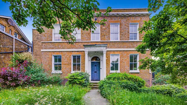 A two-story brick building with a central entrance, surrounded by lush greenery and a pathway leading to the door. - Modern 2BR Apartment 02 with Parking in Greenwich (London)