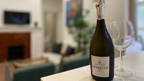 A bottle of San Vito Prosecco on a table with a glass, in a room with a fireplace and a television. - Modern 2BR Apartment 02 with Parking in Greenwich (London)