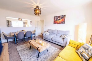 A living room with a sofa, a coffee table, a dining area with chairs, a chandelier, and a painting on the wall. - Cozy 2-Bed, 1-Bath with Free Parking, Close to Roath Park (Caerdydd)