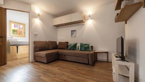 A living room with a brown sectional sofa, a small white side table with a TV, wooden flooring, and built-in cabinets. - Sunny - Cosy 1CH | 27m2 | Terrasse | 2km Remontées (Les Houches)