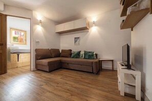 A living room with a brown sectional sofa, a small white side table with a TV, wooden flooring, and built-in cabinets. - Sunny - Cosy 1CH | 27m2 | Terrasse | 2km Remontées (Les Houches)