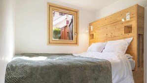 A single bed with a wooden headboard, a window with a view of buildings, and a bedside lamp. - Sunny - Cosy 1CH | 27m2 | Terrasse | 2km Remontées (Les Houches)