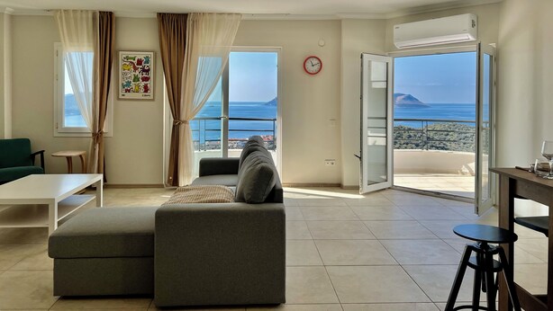 A living room with a sofa, a table, a chair, a window with curtains, a clock, and an air conditioning unit. - Lacivert Apart - Modern House with 180 Degree Unique Kaş View (Gazi)