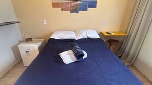 Una cama bien hecha con una colcha azul, una mesita de noche con una lámpara y una obra de arte en la pared que representa una escena de playa.
