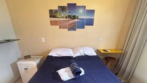 Una cama bien hecha con una colcha azul oscuro, una mesita de noche con una lámpara, un cuadro en la pared y una cortina en la ventana.