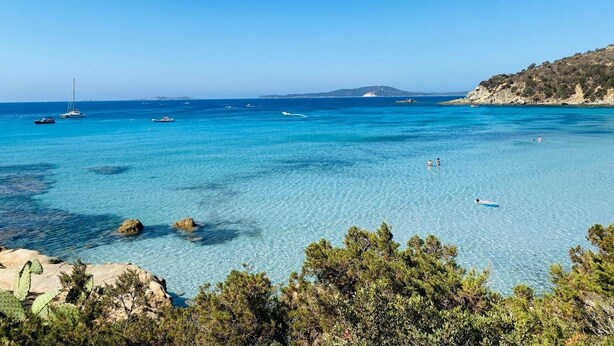 Blick auf die Küste mit klarem türkisblauem Wasser, Booten und felsigem Gelände.