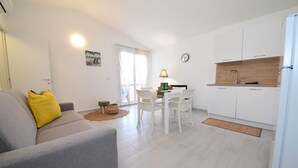 Ein modernes Wohnzimmer mit grauem Sofa, einem Esstisch mit Stühlen, einer Küchenzeile mit weißen Schränken und einer Holz Arbeitsplatte sowie einem Fenster mit durchsichtigen Vorhängen.