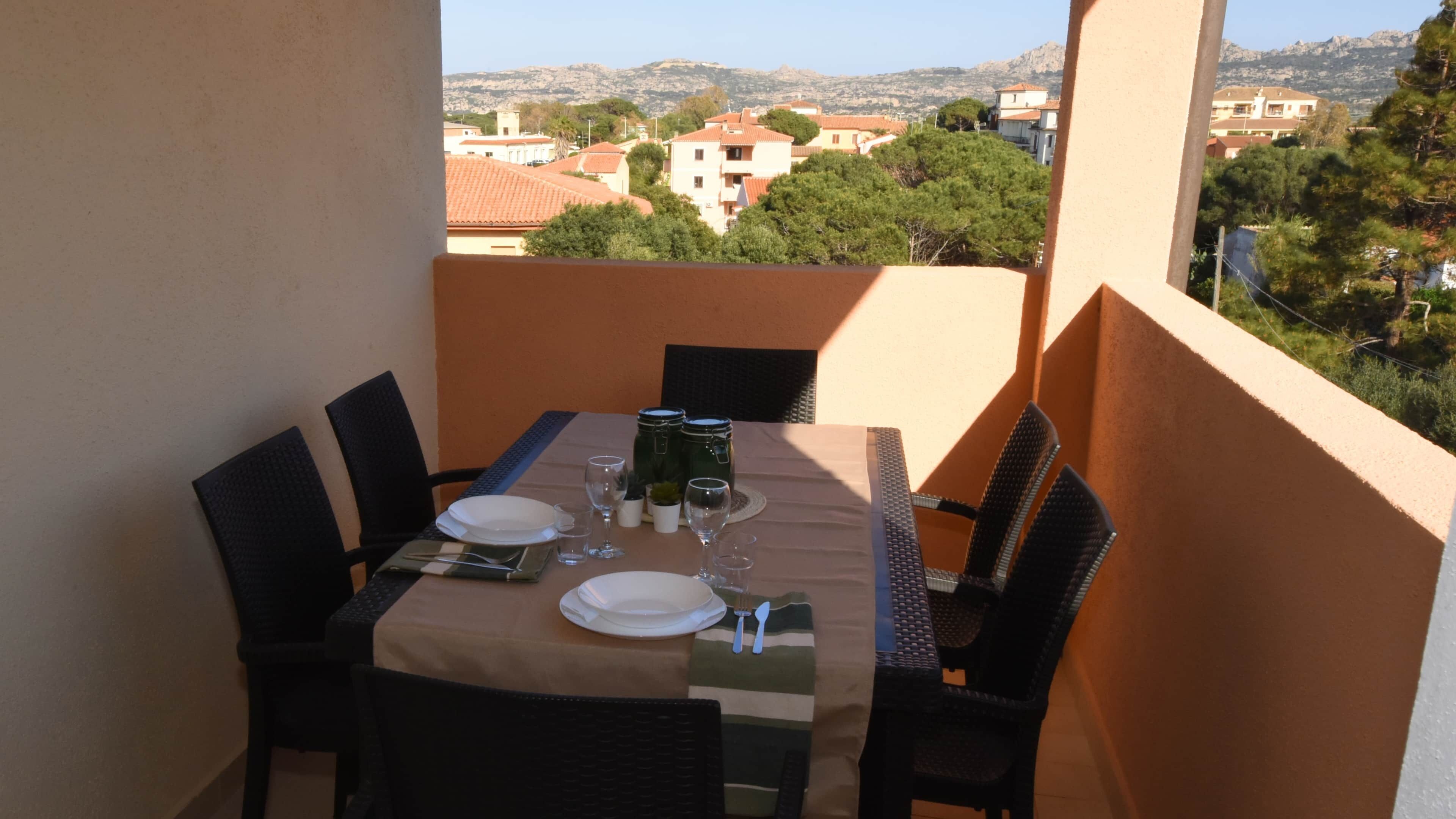 A balcony table set with plates, glasses, and cutlery, overlooking a residential area with houses and trees.