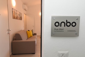 Un intérieur moderne avec un canapé beige, un coussin jaune et une enseigne fixée au mur portant l’inscription « anbo Due Mari La Maddalena ».