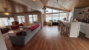 Amplia sala de estar con un ventanal que da a un lago, piso de madera y una cocina con taburetes de bar.