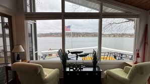 Una habitación con dos sillones verdes, techo de madera, ventanas amplias con una bandera estadounidense y vista a un lago.