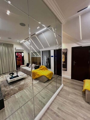 Un salon moderne avec un canapé, une table basse, un lit recouvert d’une couverture jaune, un grand miroir et une porte.