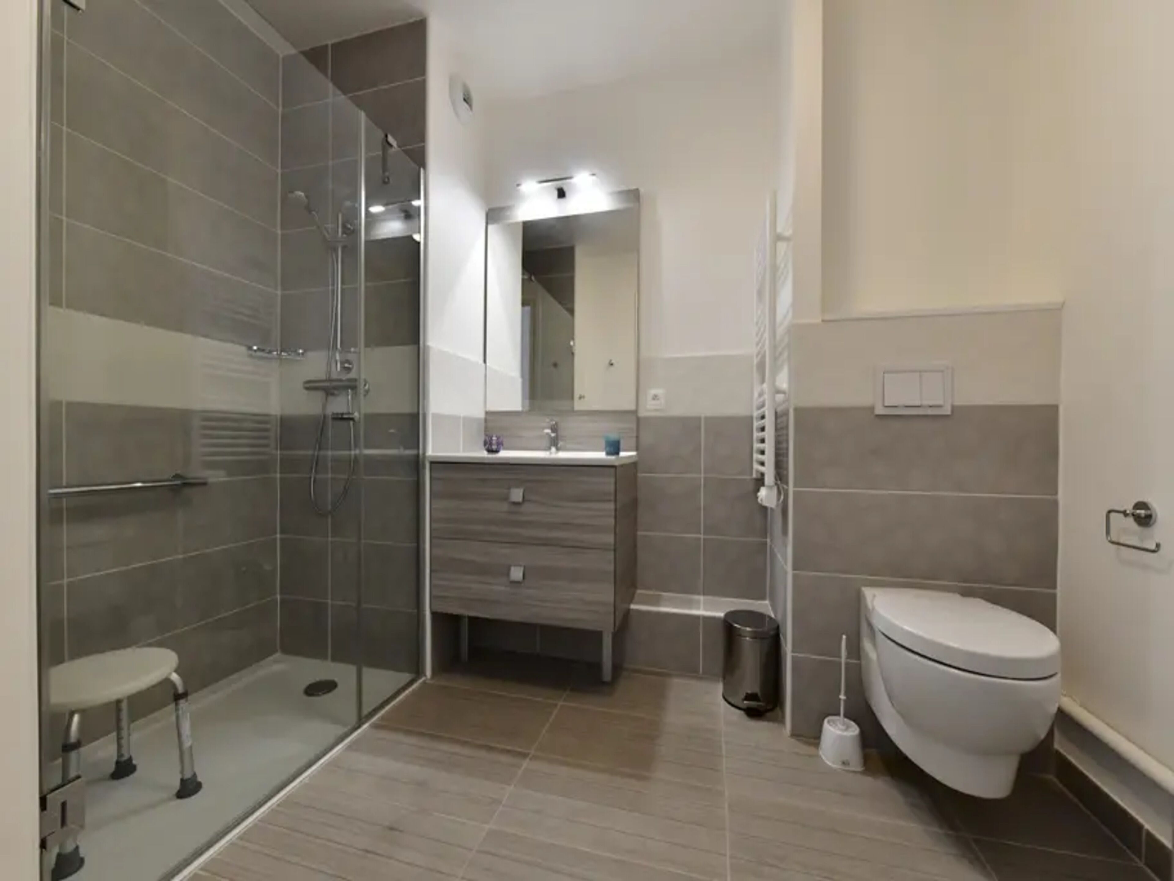 A modern bathroom with a glass shower enclosure, a toilet, a sink with a mirror, and a small trash bin.