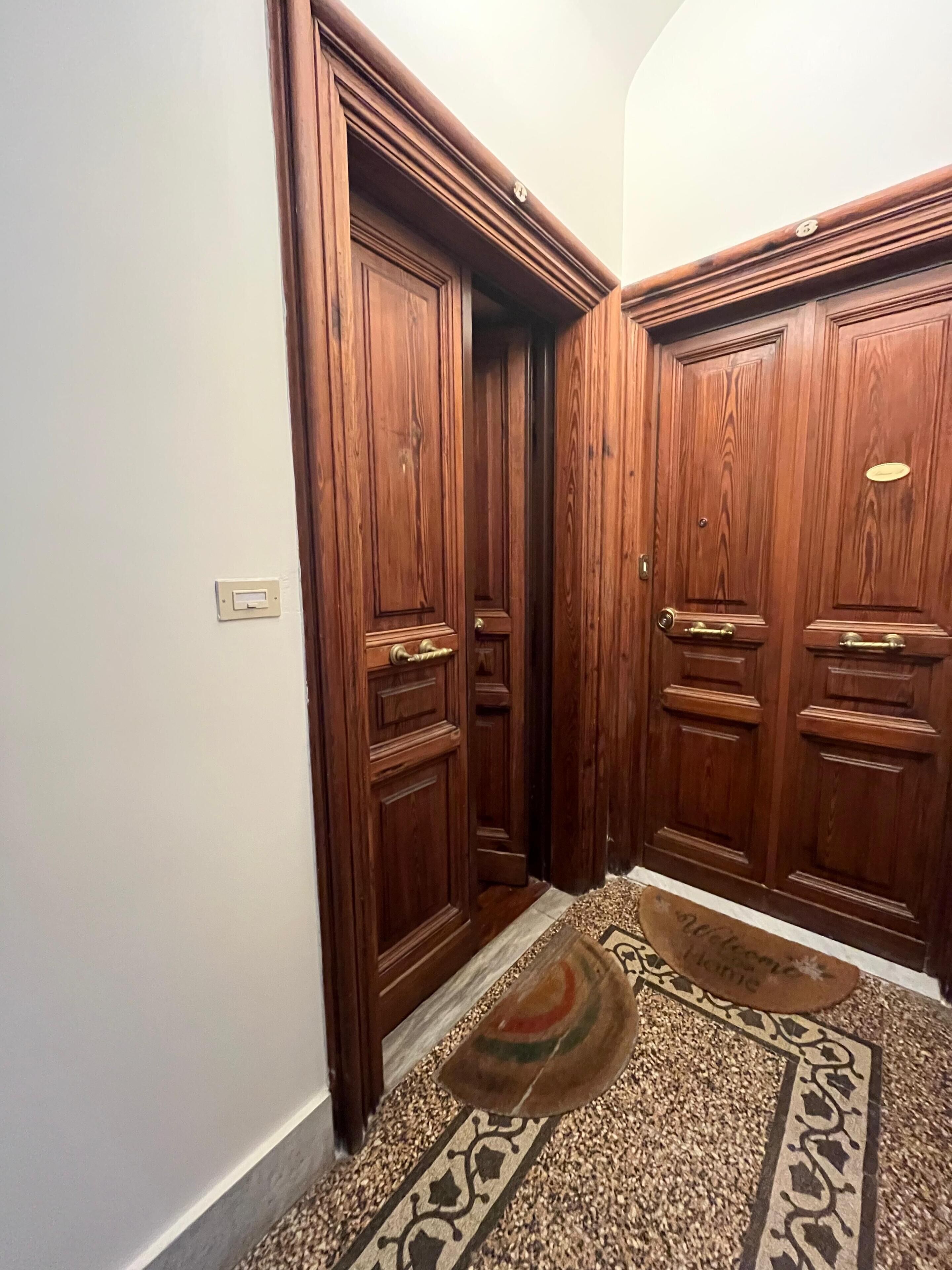 A hallway with wooden double doors, a doormat, and a light switch on the wall.
