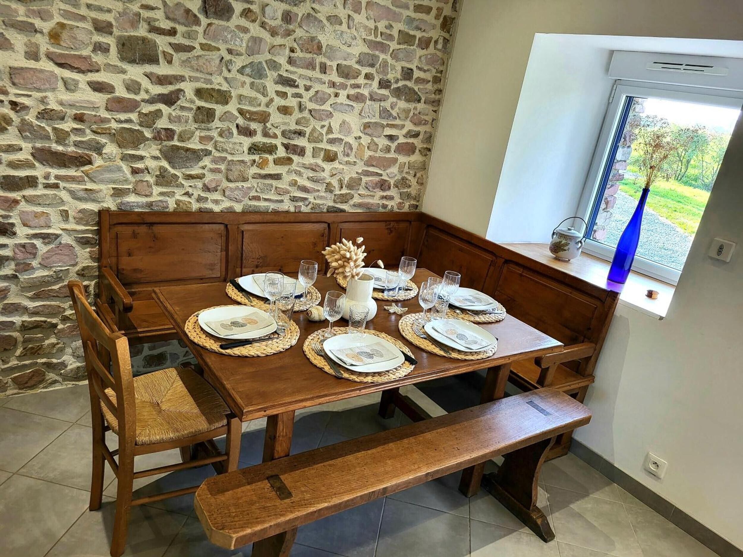 Un comedor con pared de piedra, bancos de madera, una mesa dispuesta con platos, vasos y un adorno decorativo.