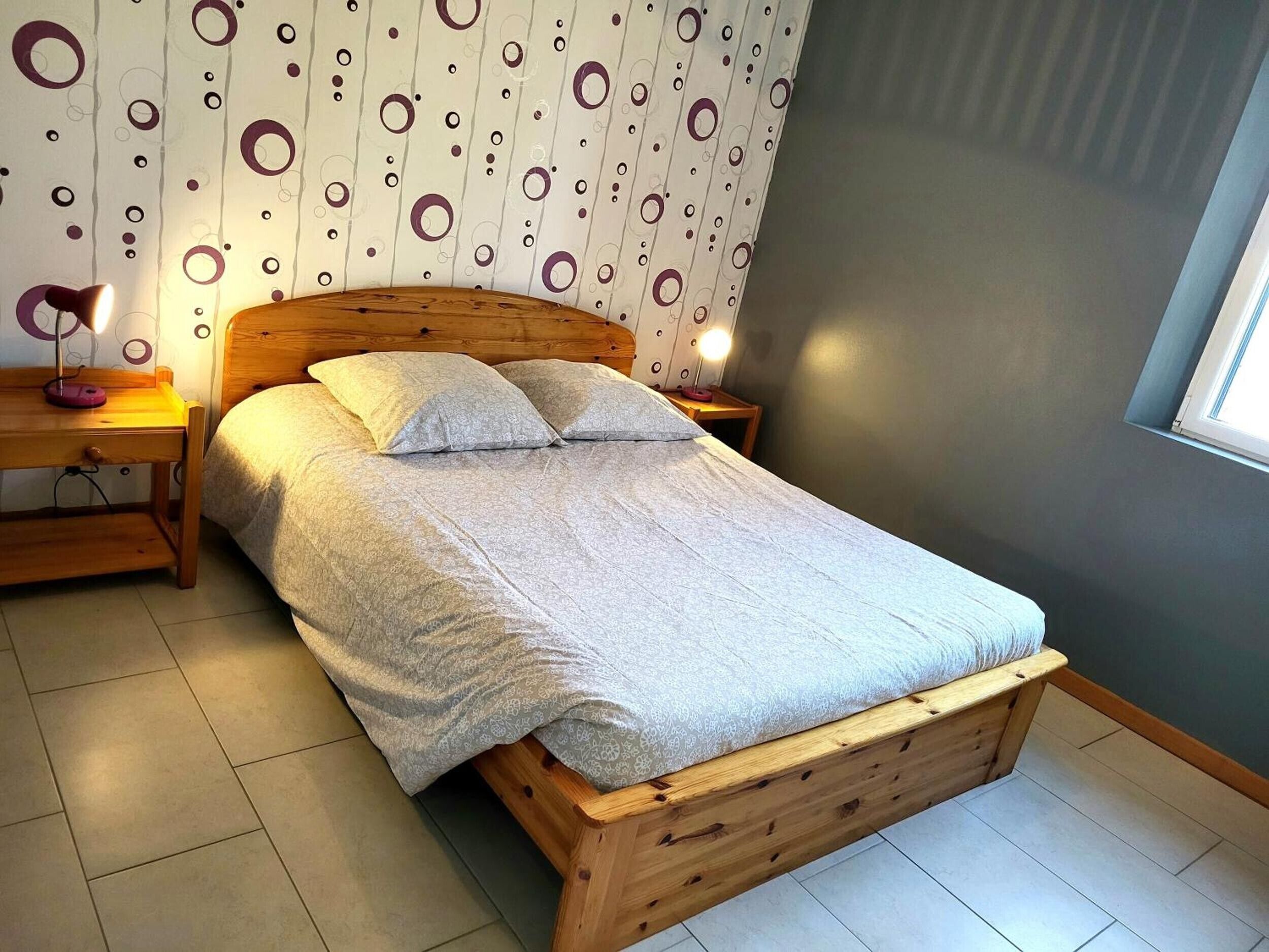 A neatly made bed with a wooden headboard, a bedside table with a lamp, and a patterned wall.