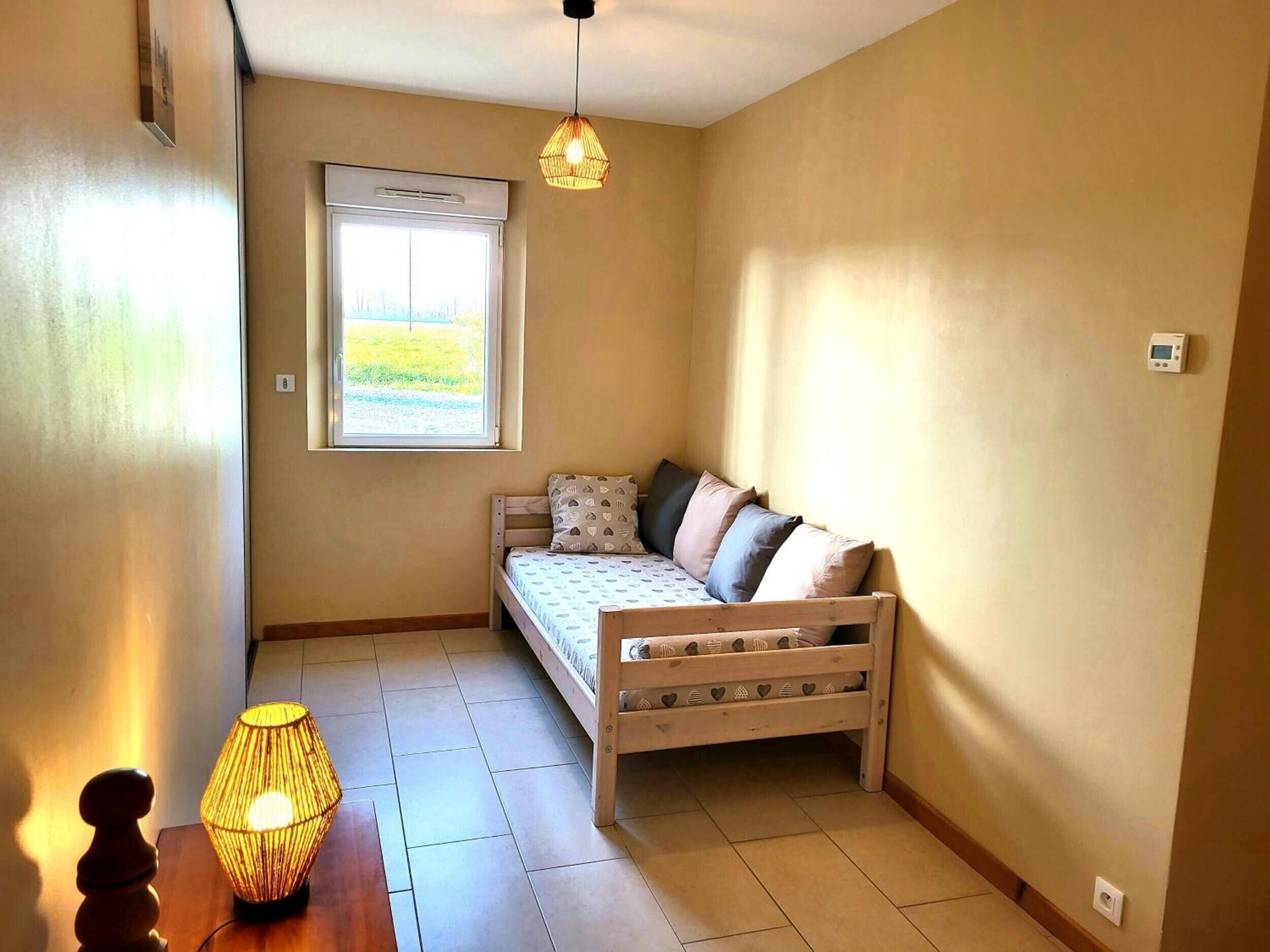 A small, cozy room with a single bed, a window with a view, a hanging light fixture, and a table with a lamp.